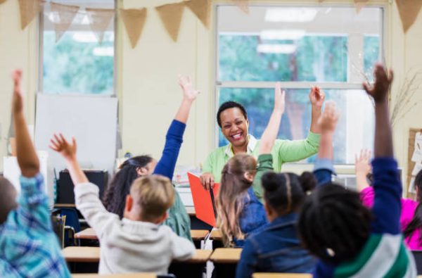 An African-American teacher, a mature woman in her 40s, sitting in front of her class of elementary school students, 6 and 7 years old, reading a book. She has asked a question and most of the children are raising their hands. They are in first grade or second grade.