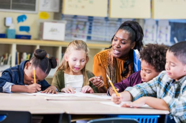 A mature African-American woman with a group of four multi-ethnic elementary school children, sitting together at a table in the classroom. The teacher is talking to her students who are writing with pencils on paper.