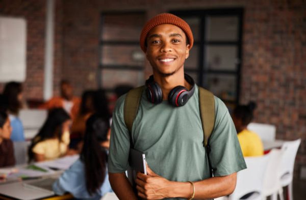 Portrait of a young male college student wearing headphones and a beanie smiling while standing in a classroom with students behind him